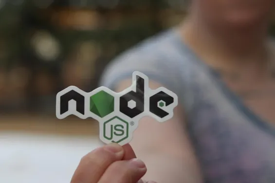 En person håller upp en klistermärke med Node.js-logotypen, som är grön och svart med en hexagonformad symbol. Bakgrunden är suddig och oskarp.